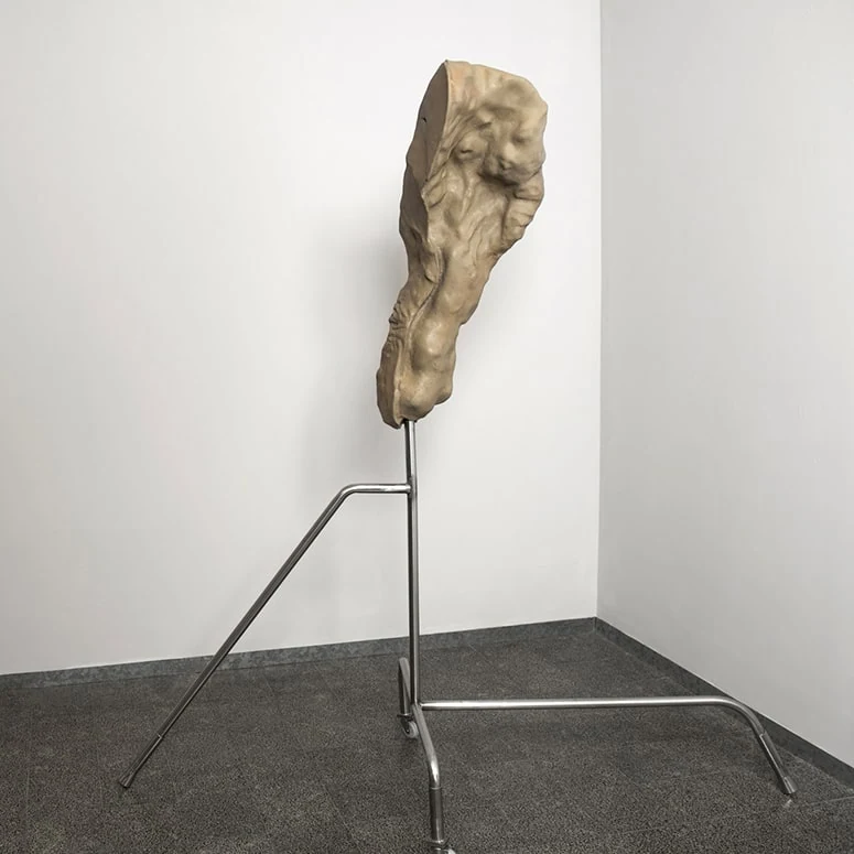 A narrow upright sculpture on a metal stand that bends outward along the gallery floor.