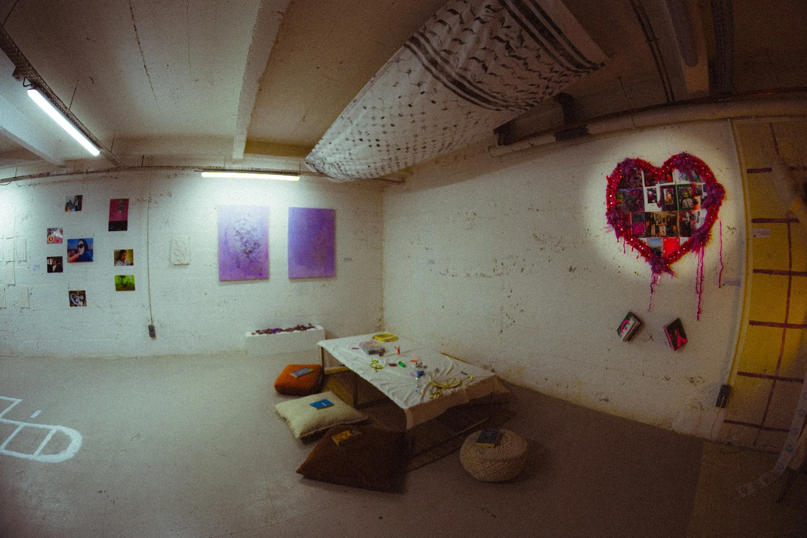 A wide view of the exhibition, with a low table, cushions, wall works and suspended fabric.