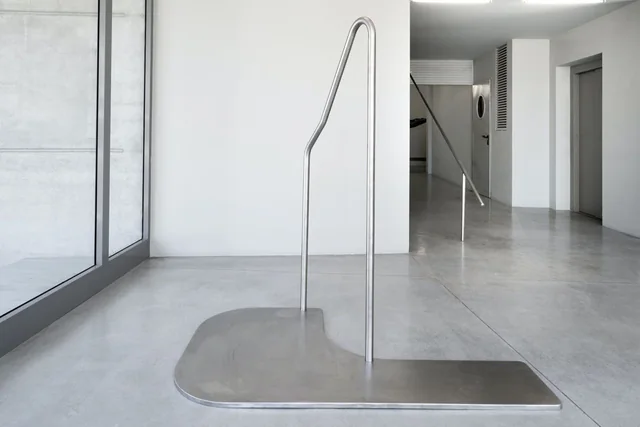 A slender metal sculpture on a curved floor plate in The Lobby Art Space.